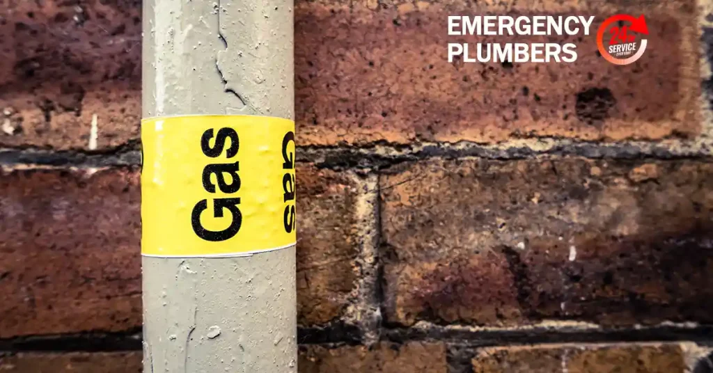 A gas pipe labeled "Gas" with a yellow sticker is mounted on a brick wall. The text "Emergency Plumbers" is visible in the upper right corner.