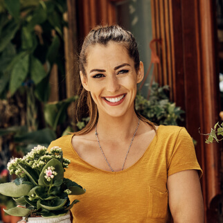 Smiling woman in a yellow shirt holds a potted plant, standing in front of a wooden door with plants visible in the background.