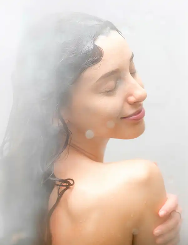 Woman with eyes closed and slight smile enjoys a shower, with visible steam and wet hair. She is shown from the shoulders up, turned slightly to the side.