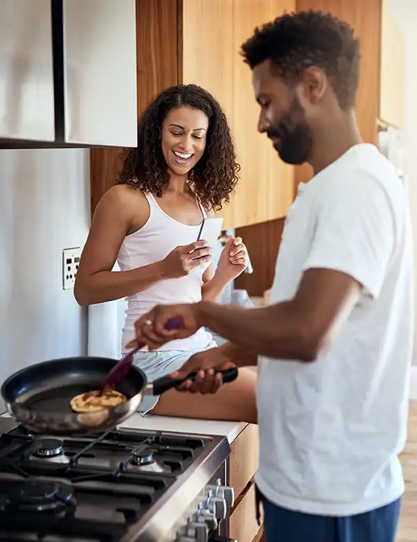 A man cooks food in a frying pan on a stove while a woman sits nearby, smiling and holding a smartphone.