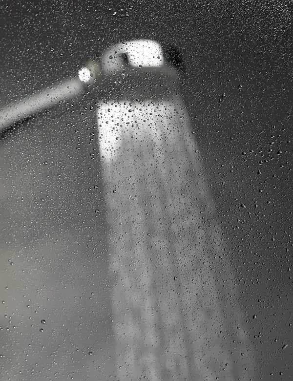 A showerhead spraying water captured from behind a glass surface covered in water droplets and steam.