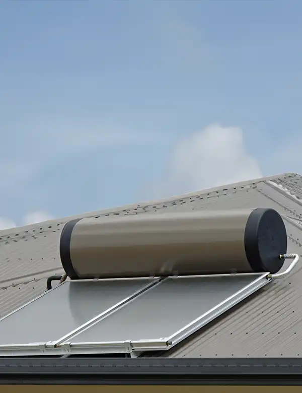 A rooftop solar water heater with a cylindrical tank and flat solar panels is mounted on a metal roof under a clear sky.