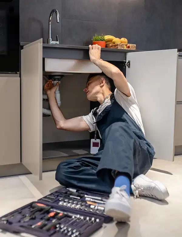 A plumber wearing overalls is working under a kitchen sink with the cabinet doors open. Various tools are spread out on the floor nearby.