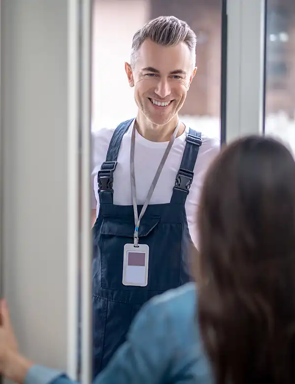 A smiling emergency plumber in overalls with an ID badge stands at a door, greeted by a female customer inside.