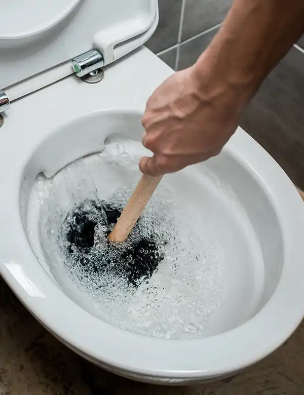 A plumber's hand uses a wooden handle plunger to unclog a flushing toilet.