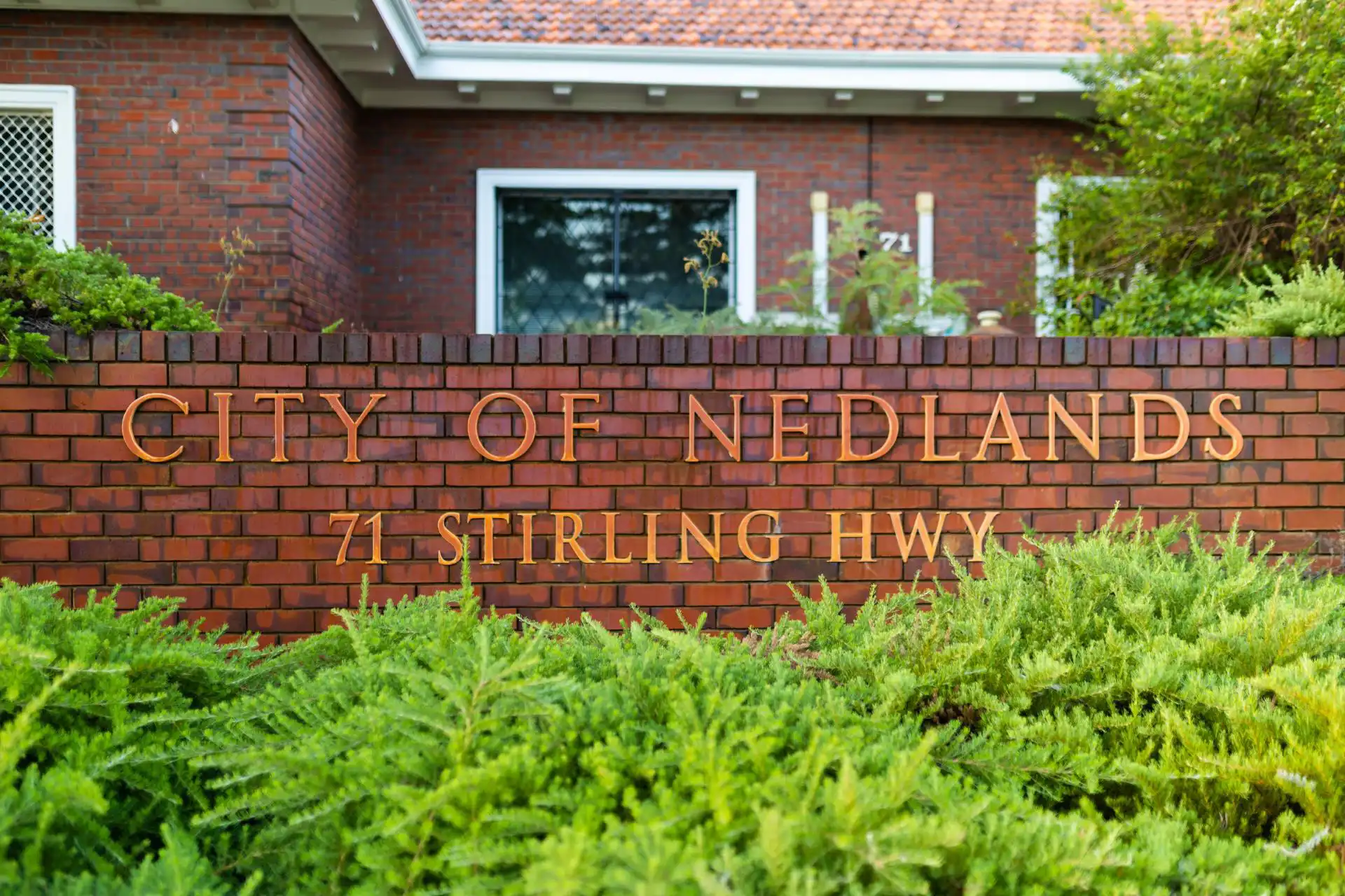 Brick sign for "City of Nedlands, 71 Stirling Hwy," in front of a red-brick building with greenery in the foreground.