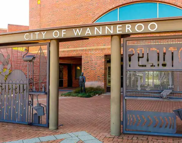 Entrance gate to the City of Wanneroo, featuring artistic metal designs, with a red brick building and a statue in the background.