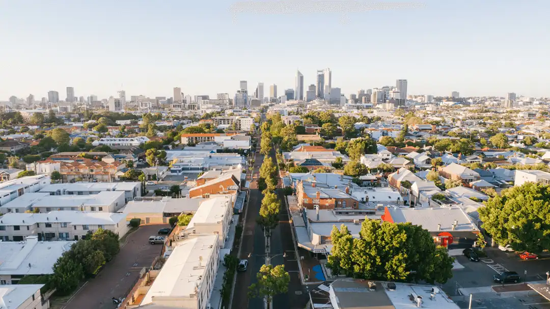 Aerial view of a residential neighborhood with tree-lined streets, leading to a city's skyline featuring several high-rise buildings under a clear, blue sky.